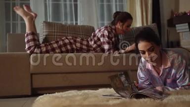 穿睡服的漂亮女孩。 一个女孩坐在沙发上时在手机上打印指纹，第二个女孩躺在地板上